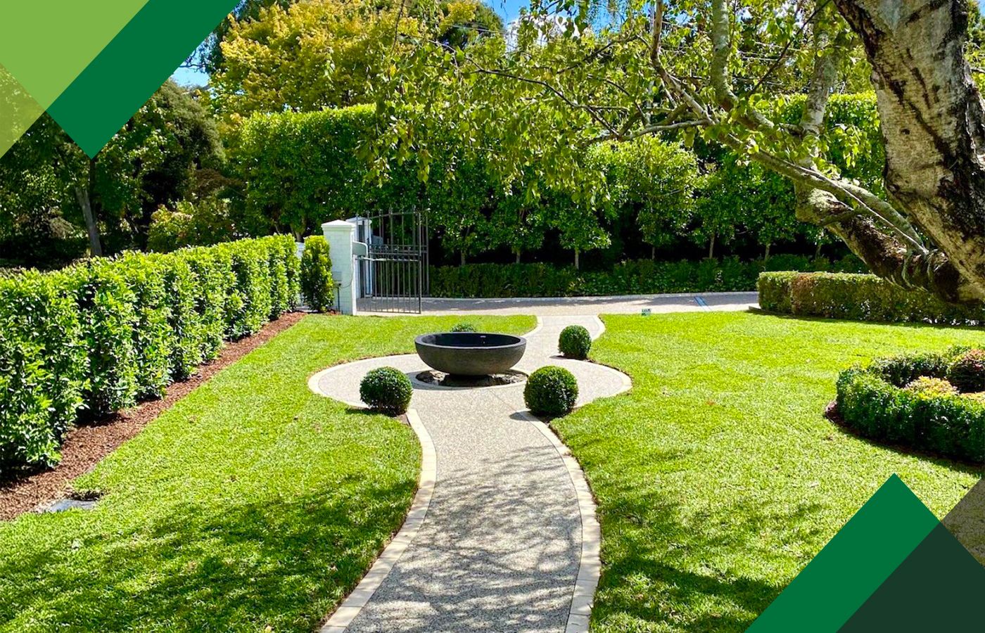 Curved stone path through a manicured garden to a round black bowl fountain, bordered by clipped hedges and trees.