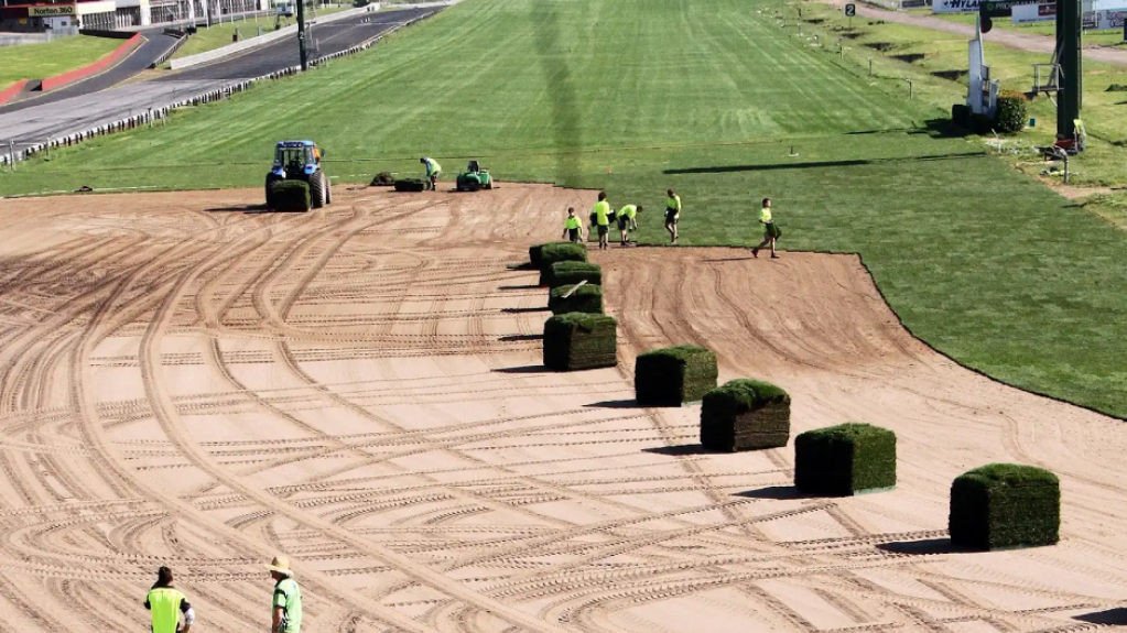 Workers laying turf across a large prepared sports field surface