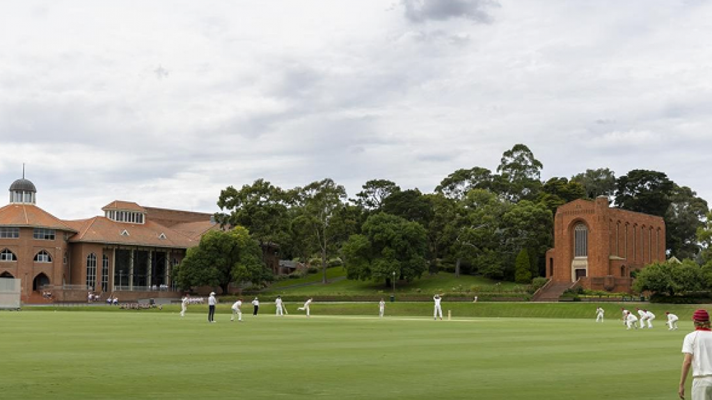 Cricket match in progress on a green oval, with players spread across the field and large brick buildings and trees in the background under a cloudy sky.