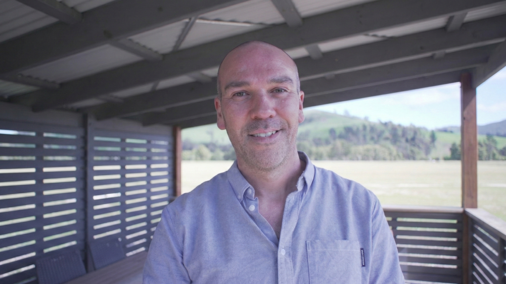 A smiling, bald man in a light blue button-down shirt standing on a covered patio with a grassy field and green hills in the background.