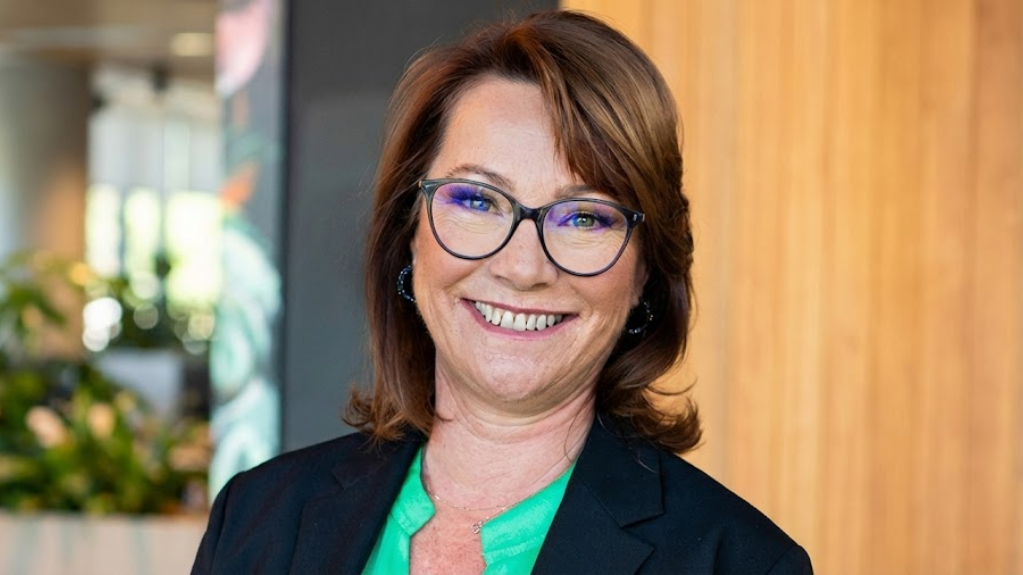 A professional portrait of a smiling woman with glasses, wearing a green top and a black blazer, with a blurred office background.