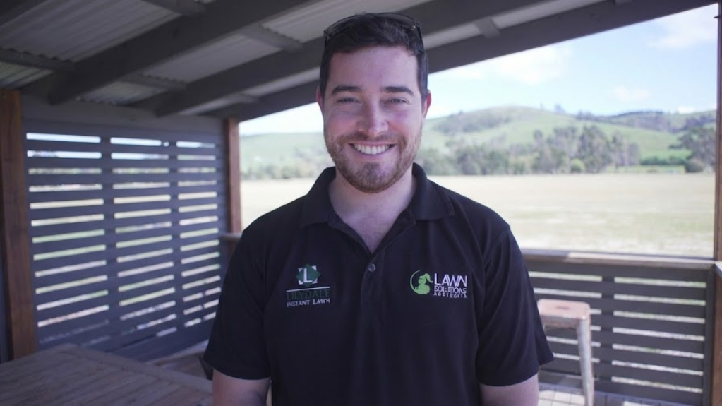 Smiling bearded man in a black logo polo standing on a covered deck overlooking a rural field.