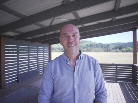 A smiling man, Andrew Batty, standing on a wooden deck with green hills in the background.