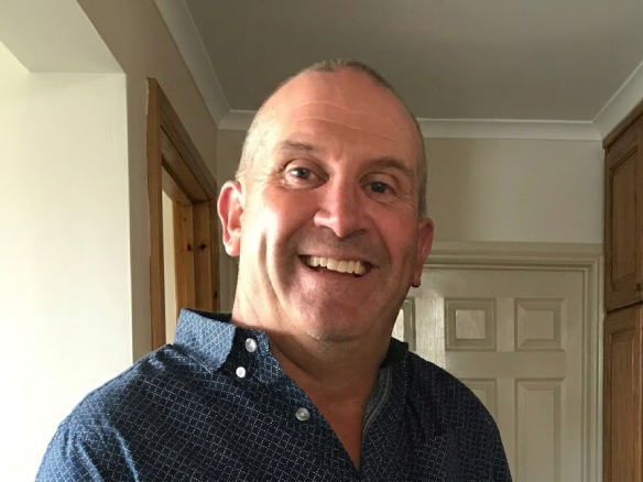 A head-and-shoulders photograph of a smiling middle-aged man with short grey hair, wearing a patterned blue button-down shirt, standing in a home kitchen environment.