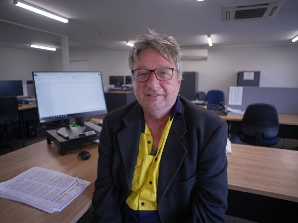 A middle-aged man with glasses, wearing a yellow high-visibility shirt under a dark blazer, sitting at a wooden desk in an office environment.