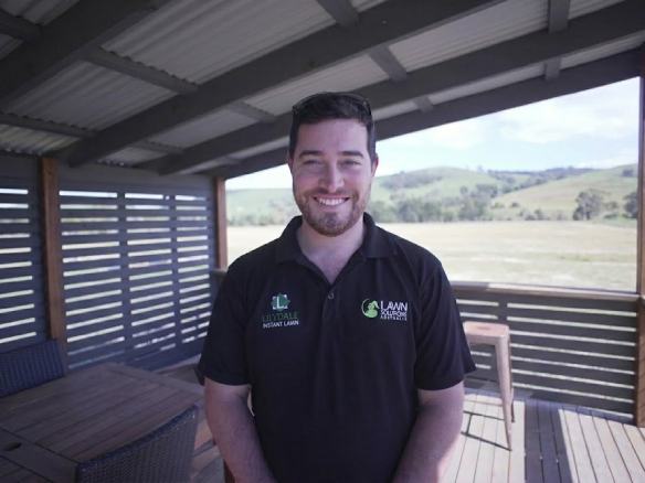 A smiling man in a black polo shirt with "Lilydale Instant Lawn" and "Lawn Solutions Australia" logos, standing on a covered deck.
