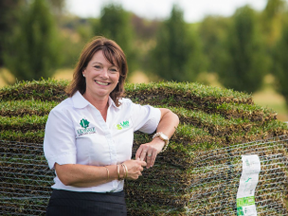 A smiling woman in a white polo shirt leaning against a large pallet of fresh grass sod at an outdoor turf farm.