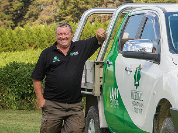 A smiling man in a black polo shirt leaning against a white Lilydale Instant Lawn delivery truck.