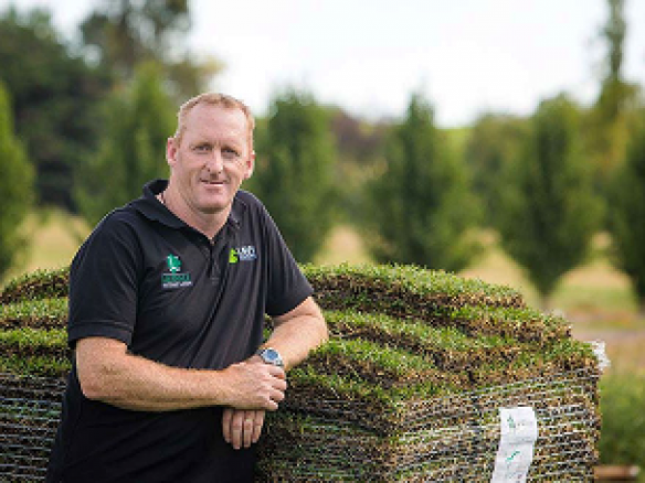 A smiling man in a black polo shirt leaning on a large pallet of fresh grass sod at an outdoor turf farm.