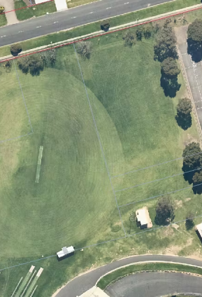 Aerial view of the Peppercorn Park Sports Field with marked cricket pitch and field lines, bordered by surrounding streets, trees, and an adjacent court complex.