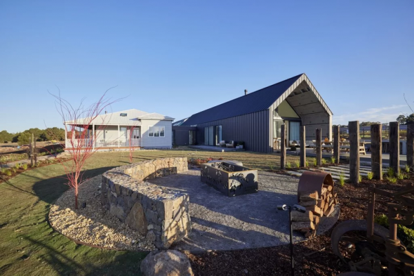 Modern rural home with a black gabled pavilion, landscaped garden beds, and a curved stone fire pit area under a clear blue sky.