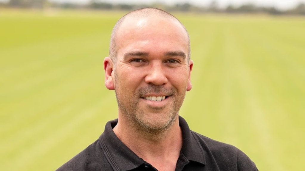 Smiling man in a black polo shirt standing outdoors with a green turf field in the background